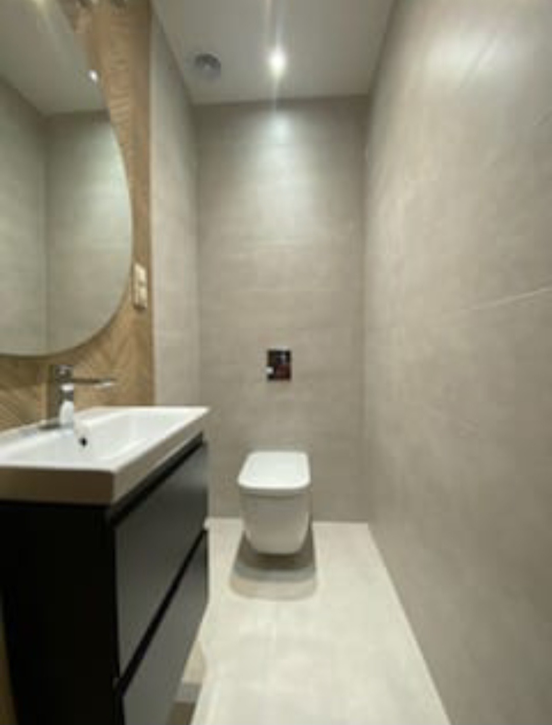 Nowoczesna łazienka w odcieniach szarości z umywalką nablatową na czarnej szafce, okrągłym lustrem z drewnianą ramą i podwieszaną toaletą.