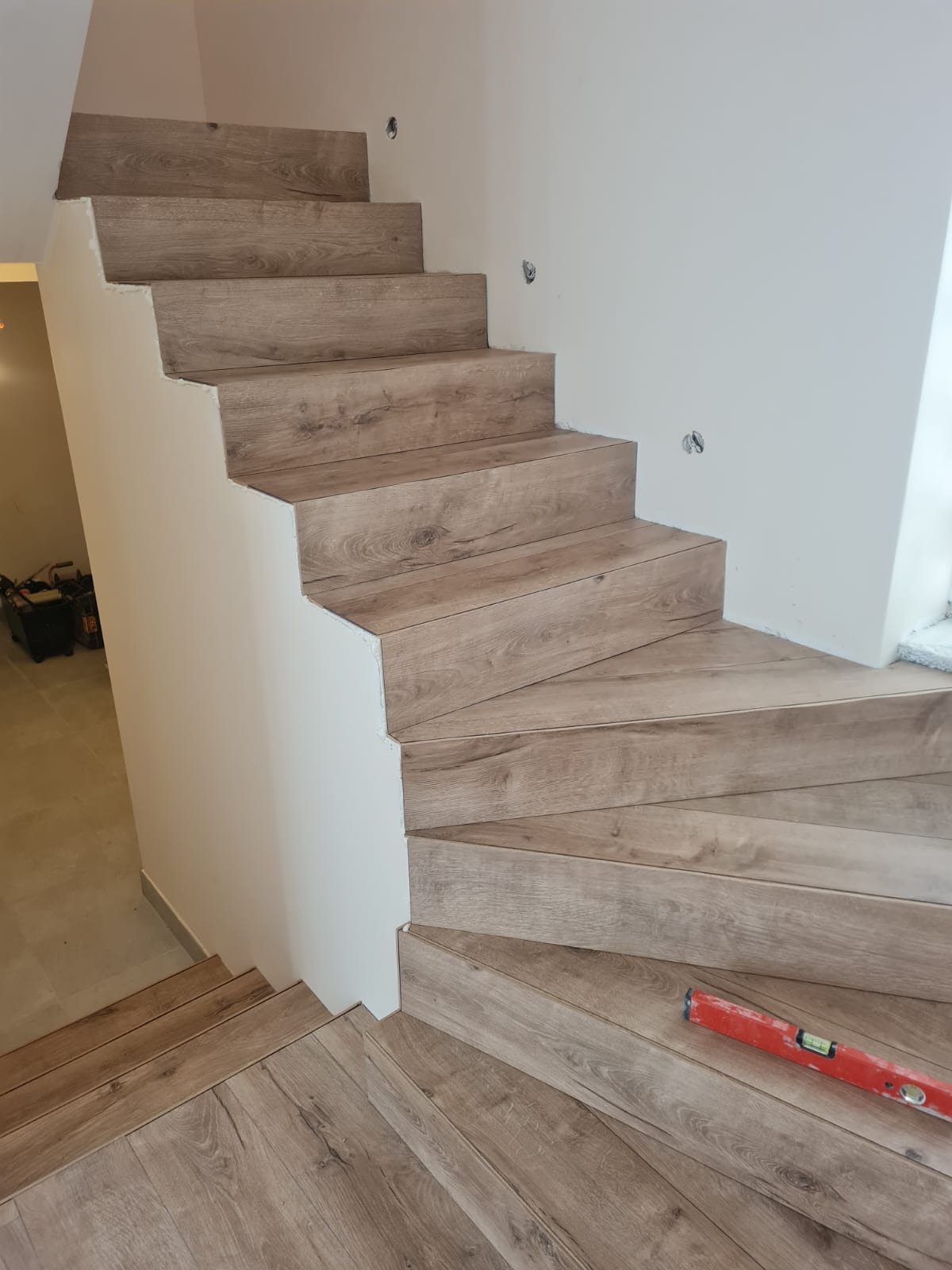 Wykończone drewnopodobne schody z białymi ścianami, widoczny poziomica na jednym ze stopni i narzędzia w tle.