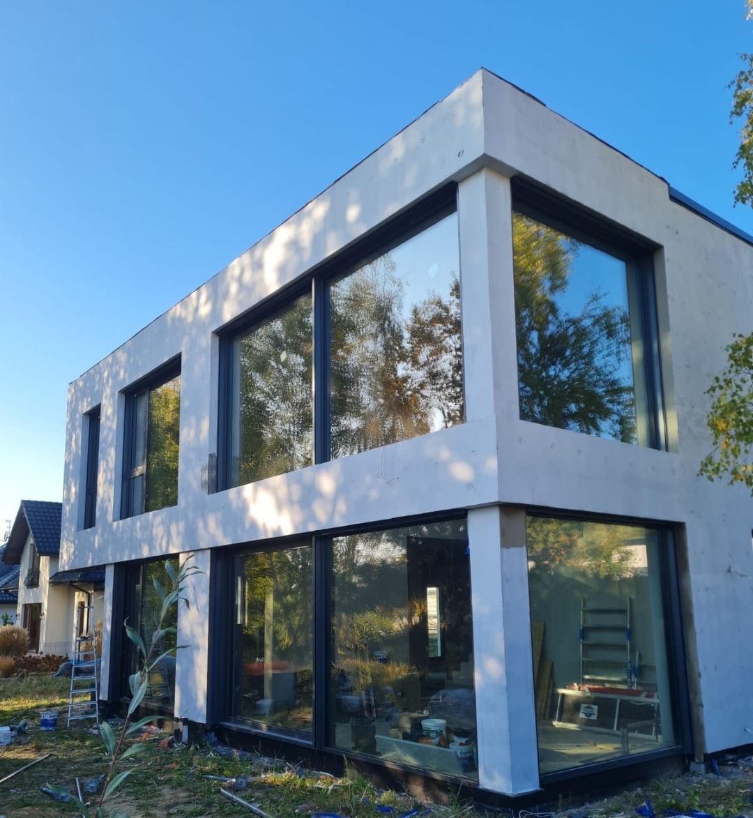 Nowoczesny dom z dużymi oknami z czarnymi ramami, elewacja w trakcie wykończenia, widoczne rusztowanie i narzędzia.