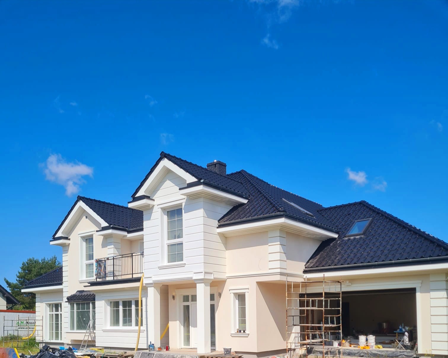 Elewacja domu jednorodzinnego w jasnych barwach z czarnym dachem, widoczne rusztowania i elementy wykończeniowe. Błękitne niebo w tle.
