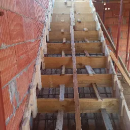 Wewnętrzne schody w trakcie budowy, drewniany szalunek zbrojony stalą, ściana z czerwonej cegły dziurawki w tle.