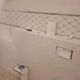 Ściana w trakcie wykańczania z białymi, wzorzystymi płytkami, widoczne narzędzia glazurnicze i otwór na oświetlenie punktowe.