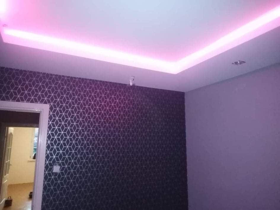 Pokój z tapetą w geometryczny wzór i podświetlanym sufitem w kolorze różowym, widoczne drzwi do innego pomieszczenia.