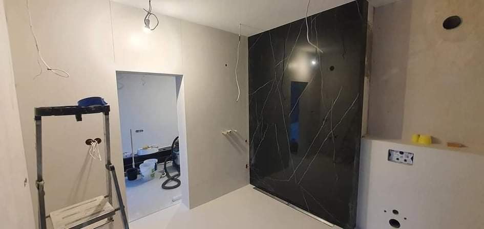 Częściowo wykończona łazienka z czarną płytą imitującą marmur na ścianie, widoczne przewody elektryczne, drabina i narzędzia w pomieszczeniu obok.