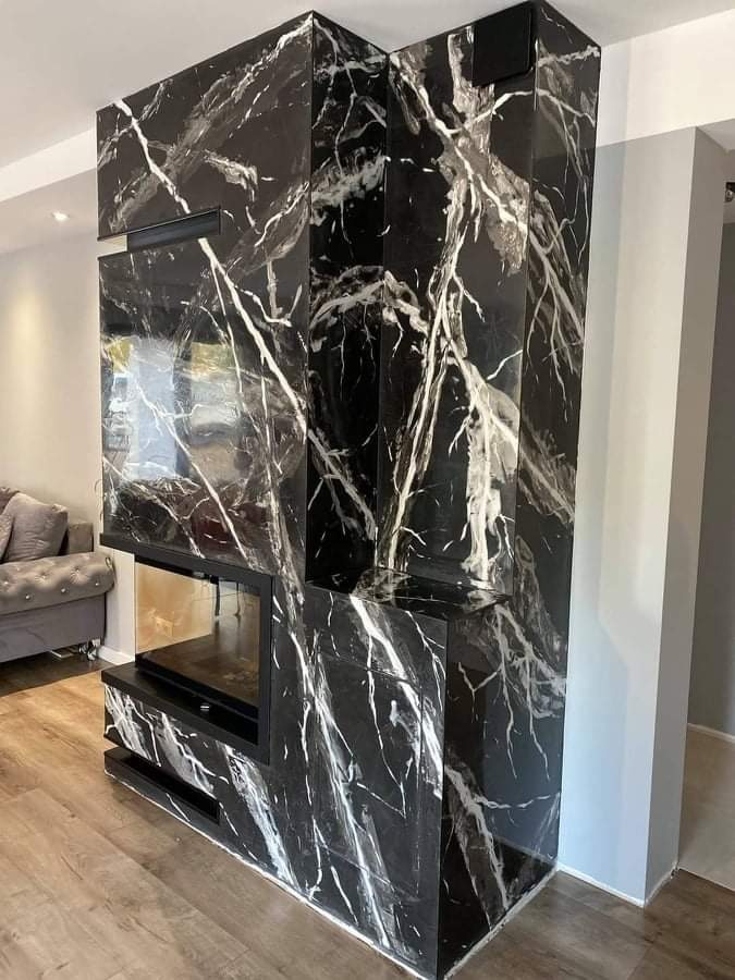 Obudowa kominka wykończona czarnym marmurem z białymi żyłami, nowoczesny design, widoczne drewniane panele podłogowe i fragment szarej sofy w tle.