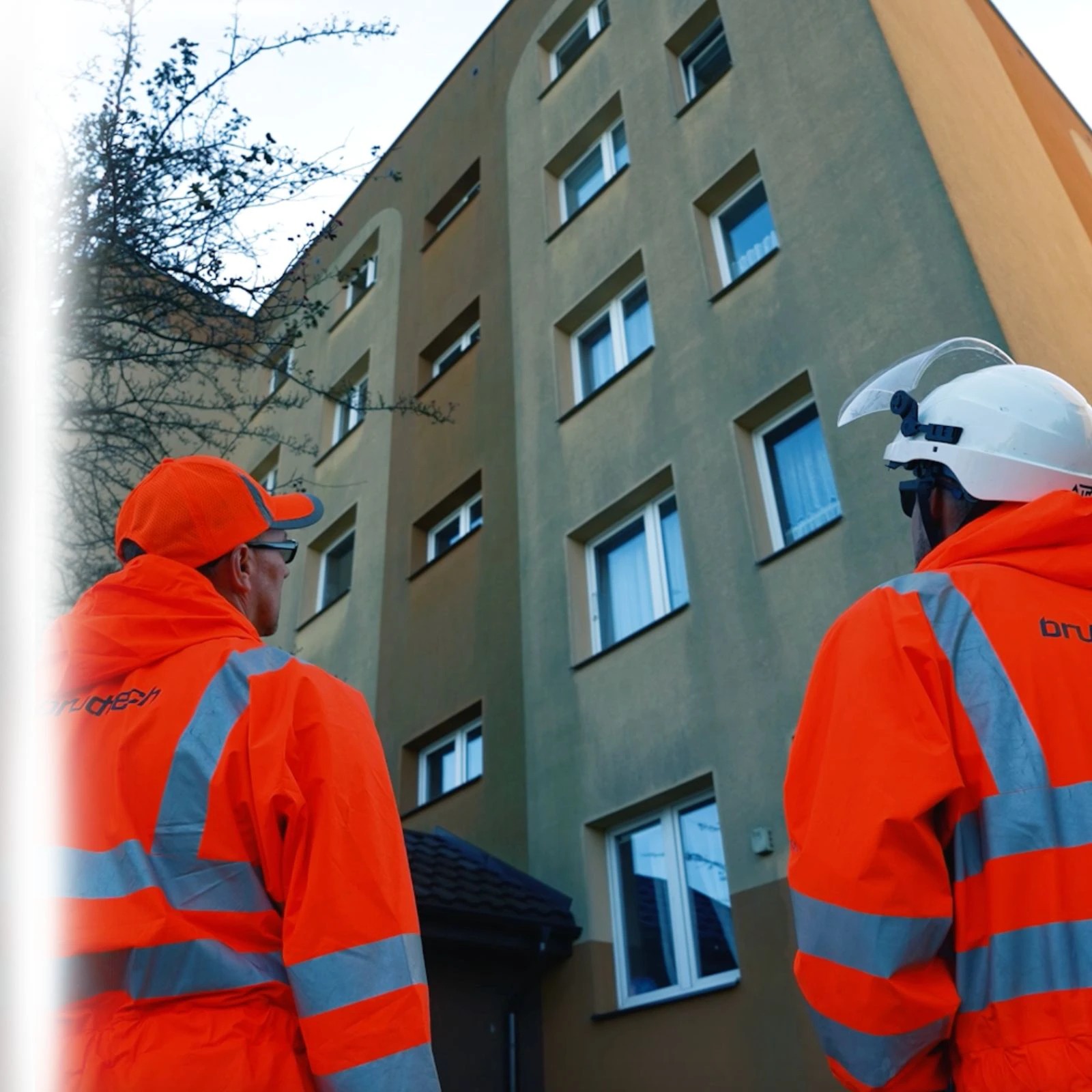 Dwóch pracowników w pomarańczowych kombinezonach i kaskach ogląda elewację budynku wielorodzinnego przed rozpoczęciem prac.