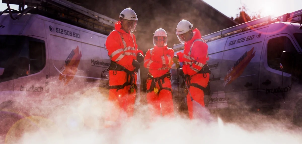 Trzech pracowników w pomarańczowych kombinezonach, kaskach i osłonach twarzy, w trakcie pracy na zewnątrz, z samochodem dostawczym firmy w tle, na którym widnieje napis 'Mycie elewacji' i 'Mycie...