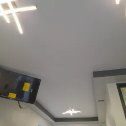 Nowoczesne oświetlenie sufitowe LED w kształcie krzyża, z widocznym telewizorem na ścianie i oknem w tle.