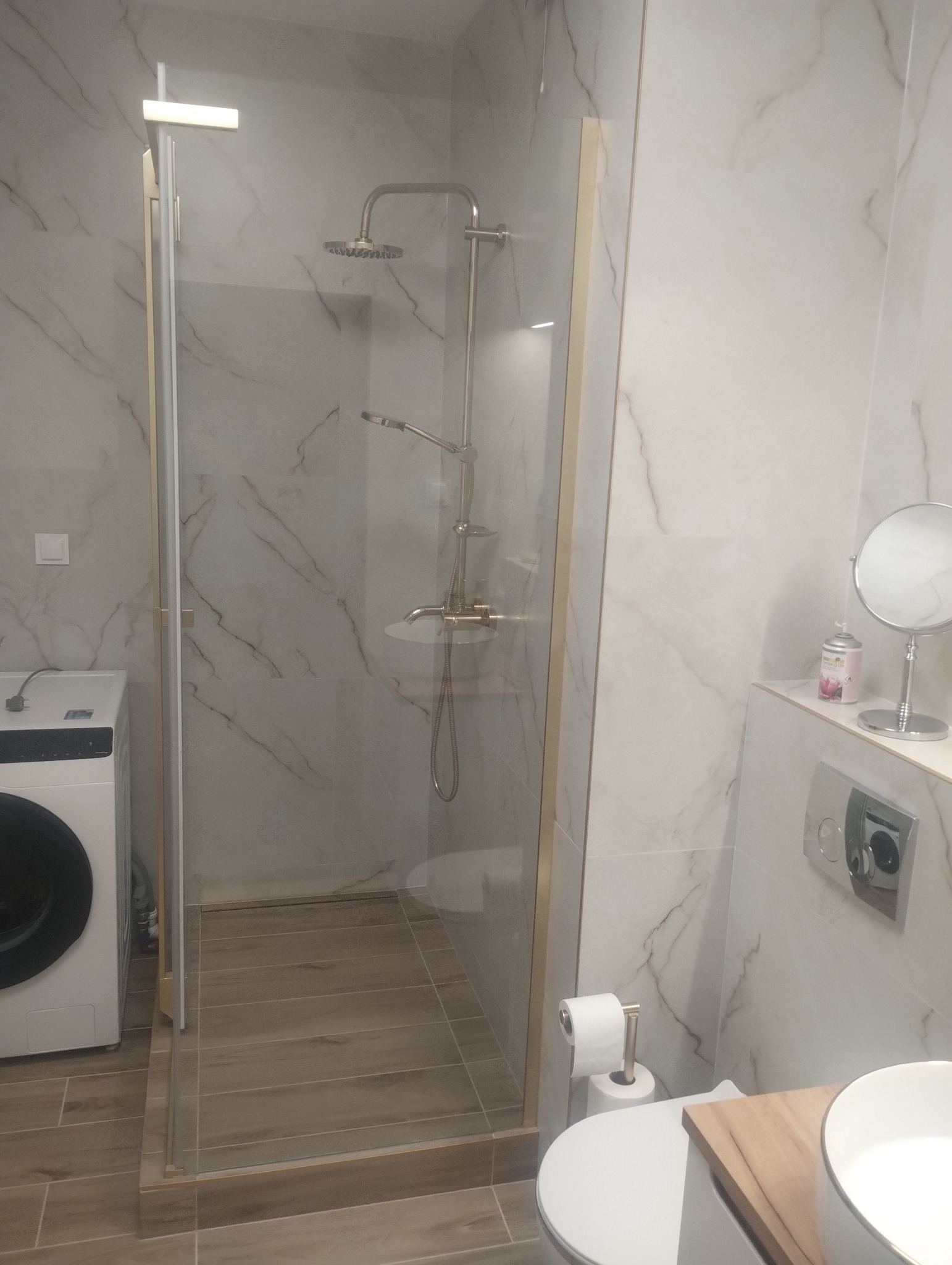 Nowoczesna kabina prysznicowa ze złotymi detalami, marmurowe płytki na ścianach, widoczna pralka i fragment toalety. Minimalistyczny design łazienki.