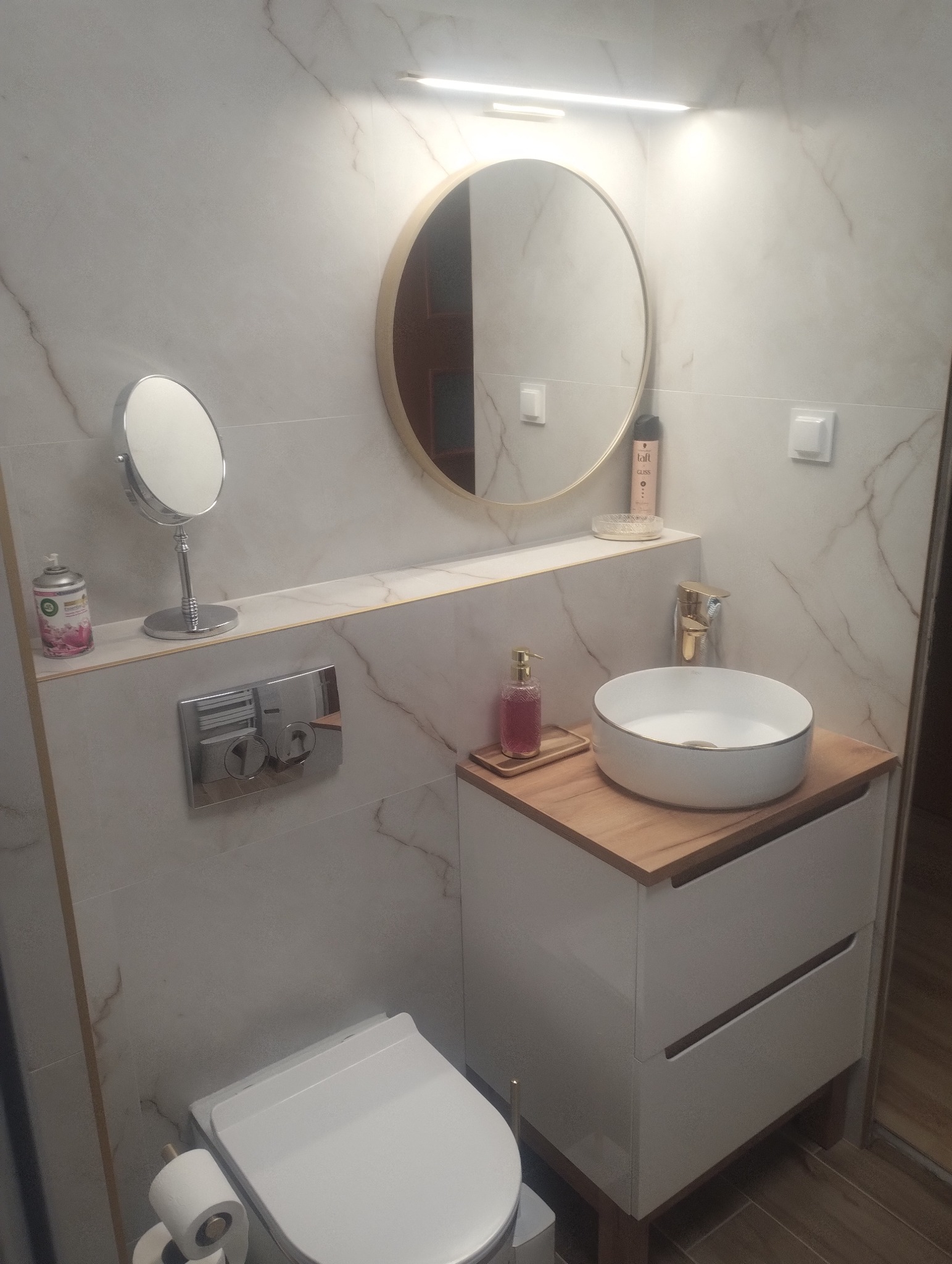 Nowoczesna łazienka z marmurowymi płytkami, okrągłym lustrem z podświetleniem LED, białą umywalką nablatową i podwieszaną toaletą. Minimalistyczny design.