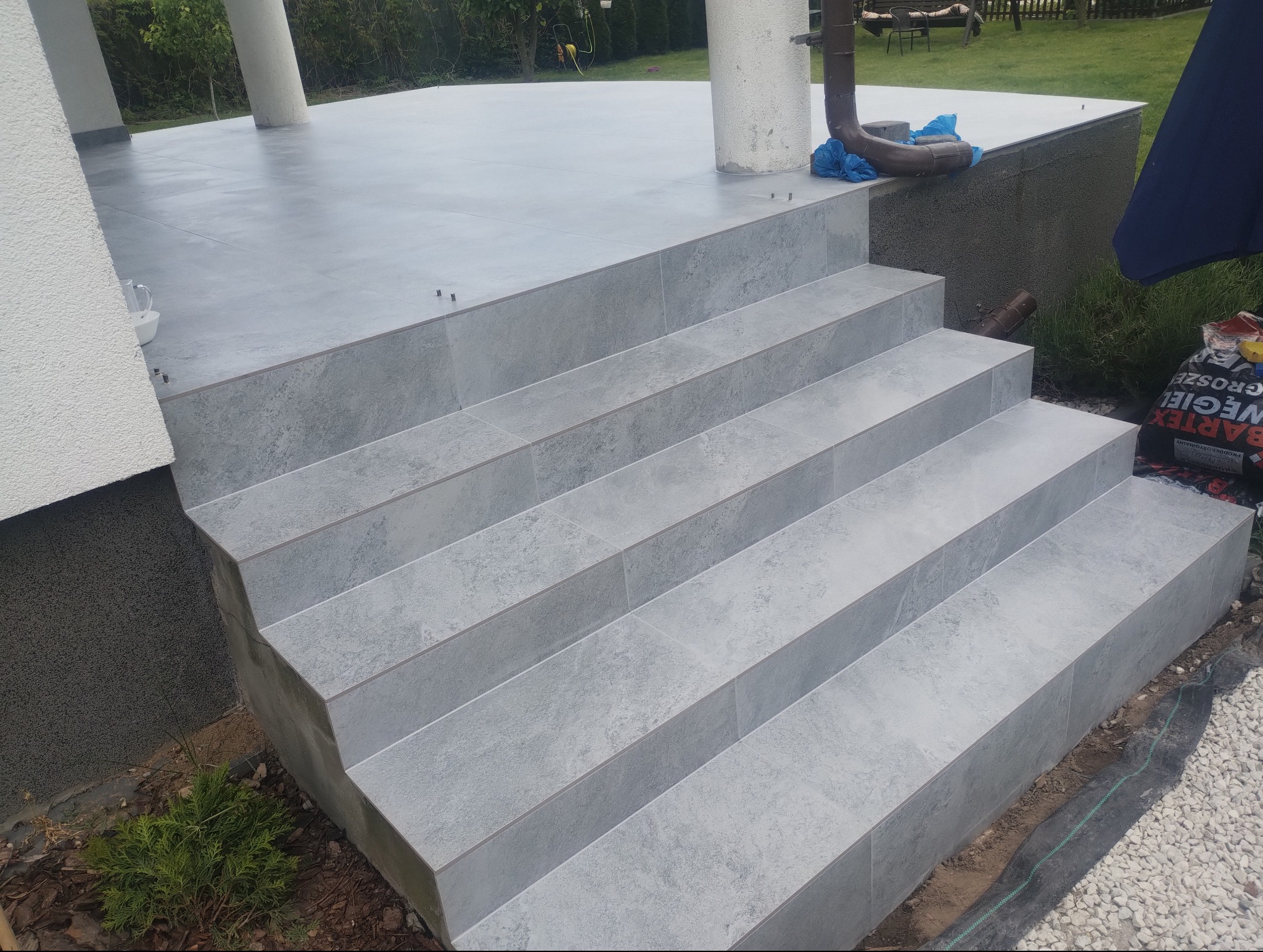 Wykończone płytkami schody wejściowe do domu, z widocznymi detalami fug i fakturą płytek. Tło stanowi trawnik i fragment elewacji budynku.