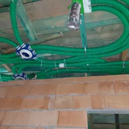 Zielone, karbowane rury wentylacyjne zamontowane pod drewnianym stropem, z widocznymi elementami mocującymi i rozdzielaczami, nad surową ścianą z cegły.