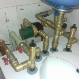 Węzeł hydrauliczny z pompą obiegową, zaworami i rurami miedzianymi, instalacja zamontowana na białej obudowie, na ścianie widoczne oznaczenia wymiarowe.