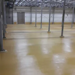 Jasnobrązowa, błyszcząca podłoga w hali przemysłowej z metalowymi słupami i systemem rur pod sufitem.