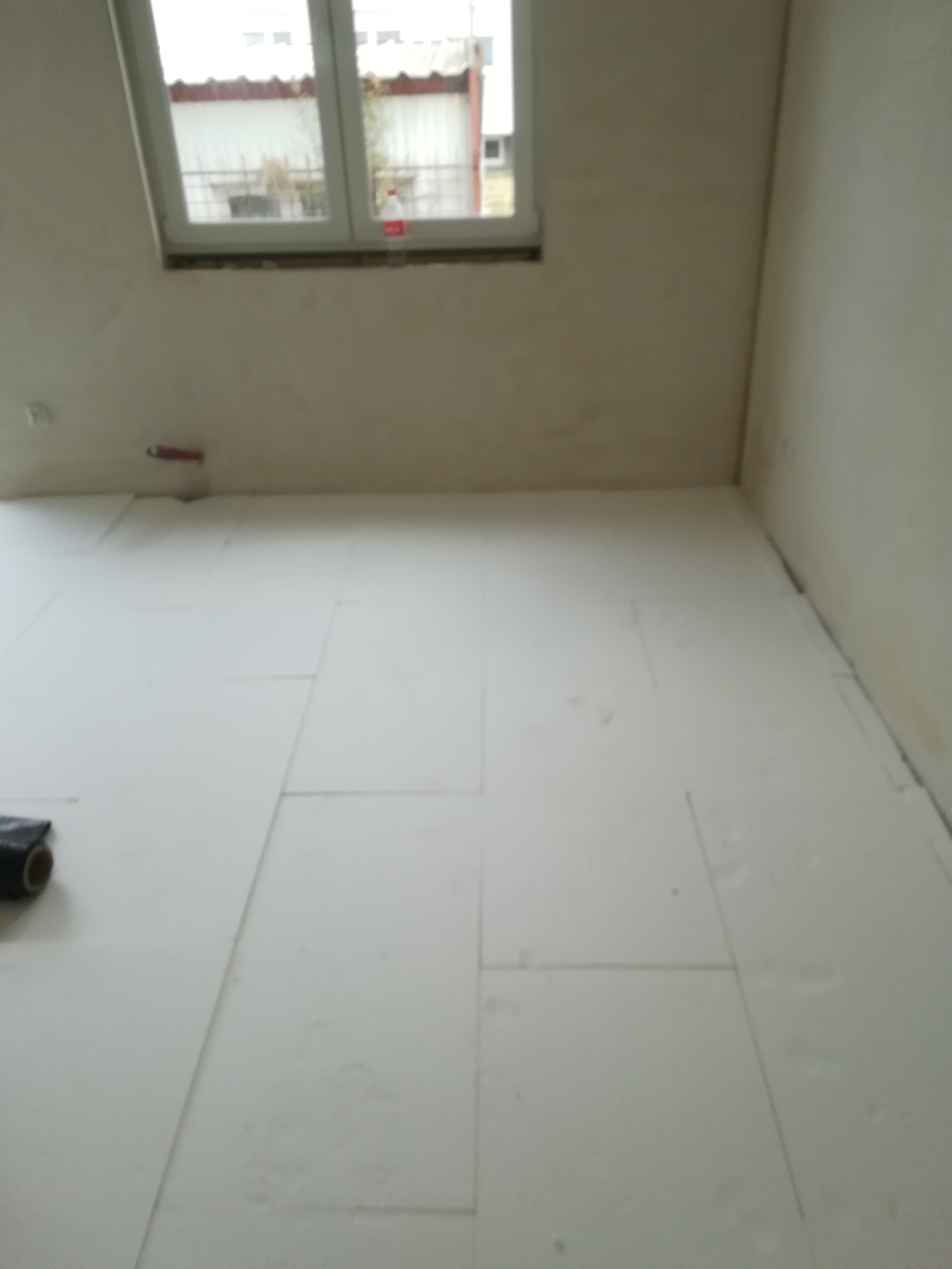 Przygotowanie podłoża pod wylewkę: białe płyty styropianowe ułożone na podłodze w surowym wnętrzu, widoczne okno i fragment rolki czarnej folii.