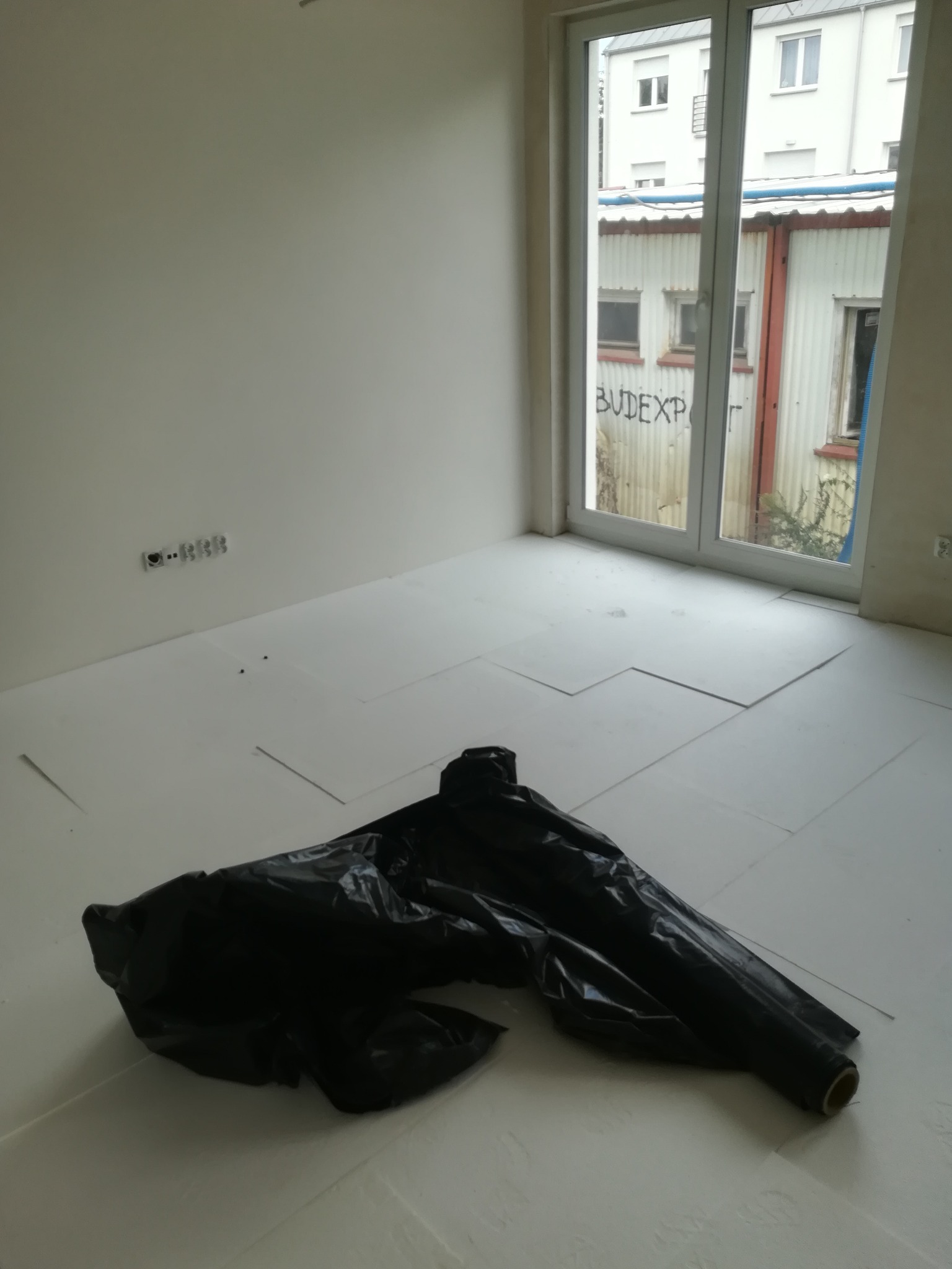 Przygotowanie do wylewki: rozłożona izolacja podłogowa z folii i arkuszy styropianu w pustym pomieszczeniu z oknem na budynek gospodarczy z graffiti.