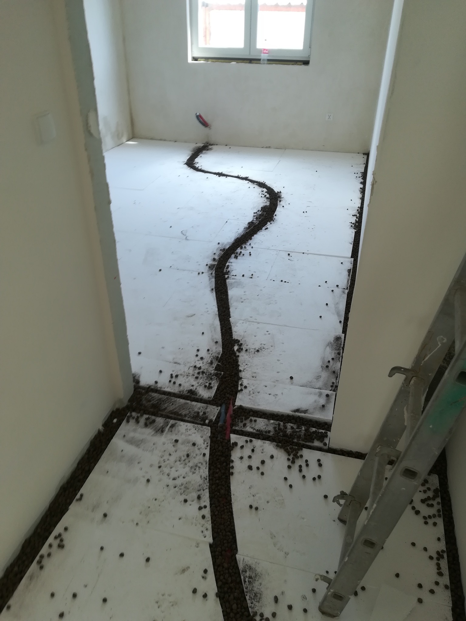 Przygotowanie podłoża pod wylewkę: rozłożony styropian i podsypka wyrównująca w pomieszczeniu z oknem, widoczna drabina oparta o ścianę.