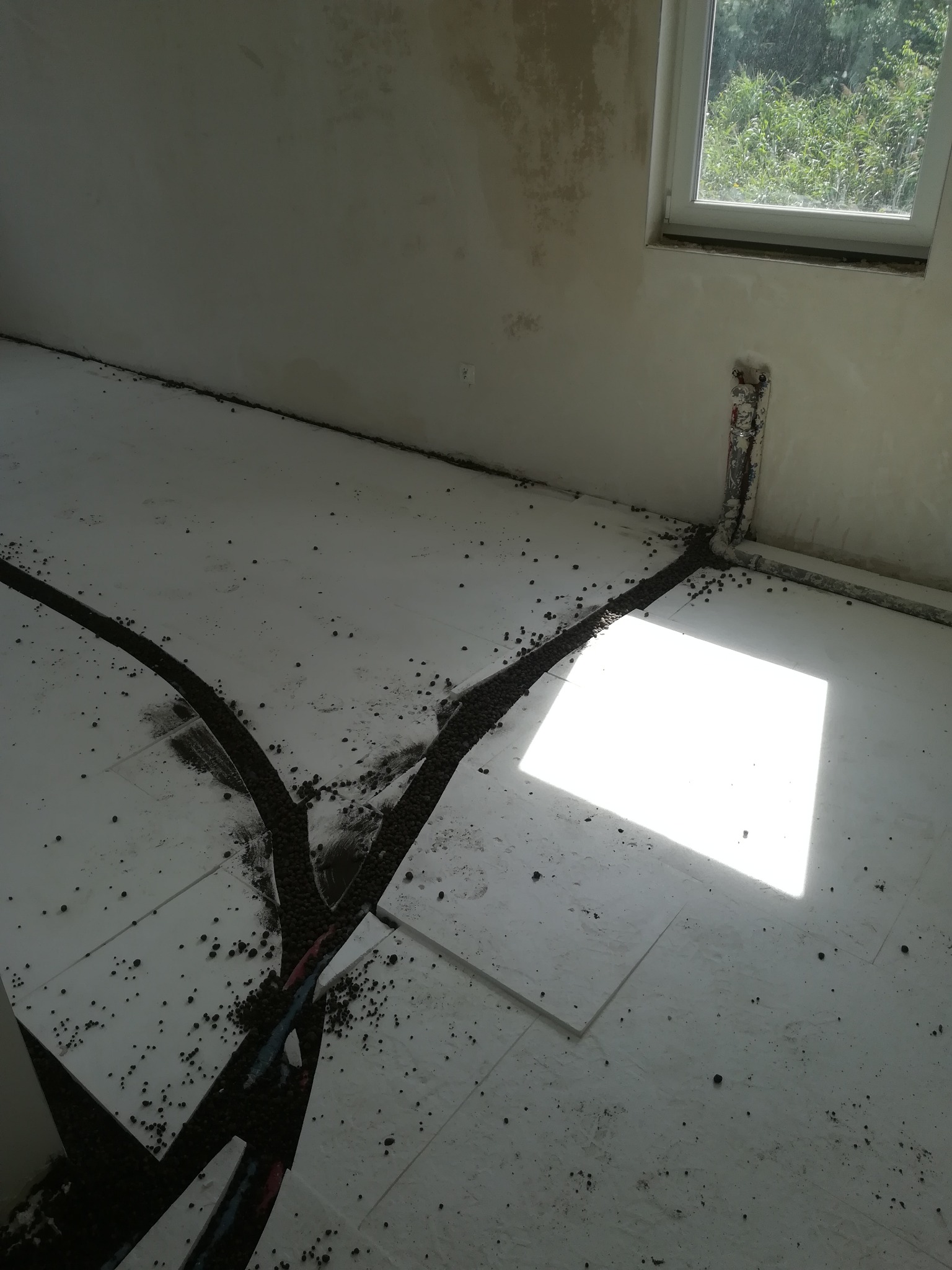 Przygotowanie pod wylewkę jastrychu: styropianowe płyty izolacyjne z widocznymi rurami ogrzewania podłogowego, zalane częściowo ciemnym, drobnym kruszywem, jasne światło słoneczne z okna.