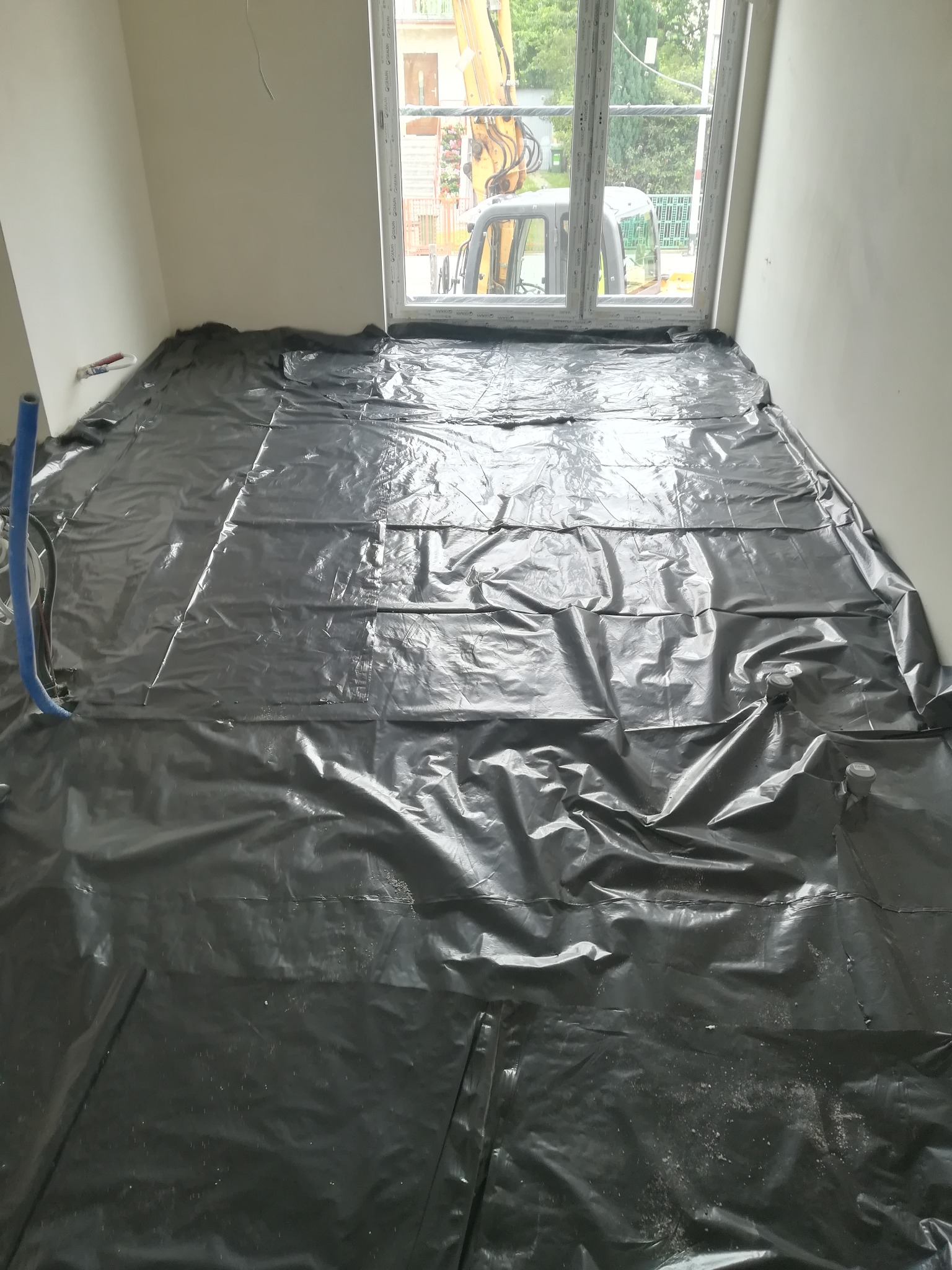 Przygotowanie podłoża pod wylewkę: rozłożona czarna folia budowlana na podłodze w pomieszczeniu z oknem, widoczne niebieskie rury przy ścianie.