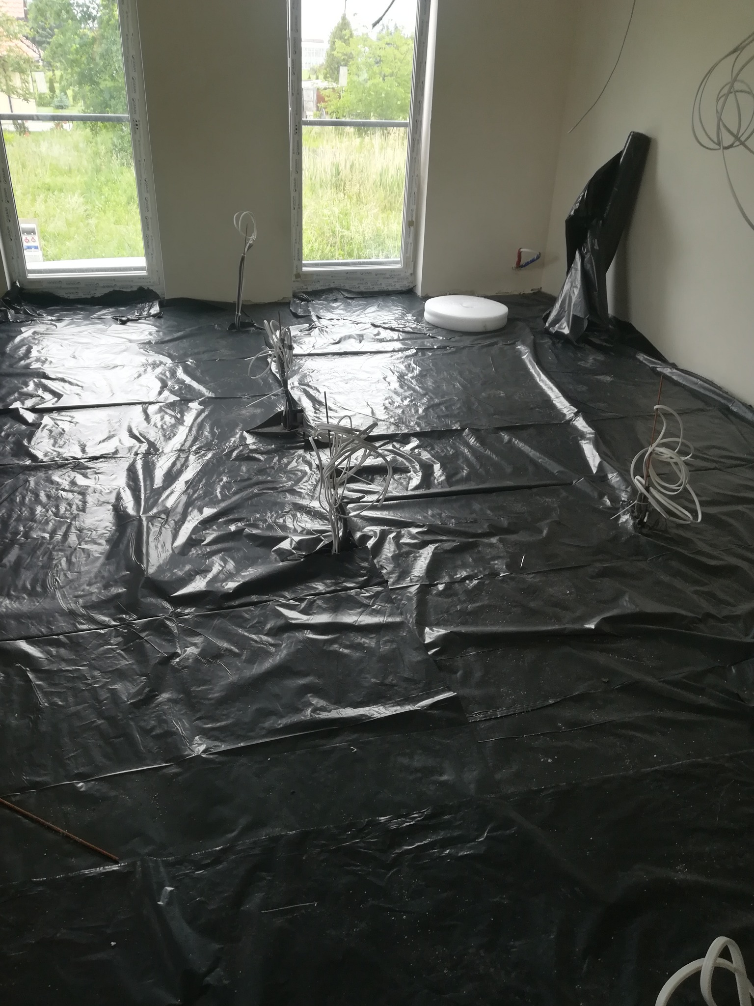 Przygotowanie podłoża pod wylewkę: rozłożona czarna folia budowlana w jasnym pomieszczeniu z oknami i wystającymi kablami elektrycznymi.