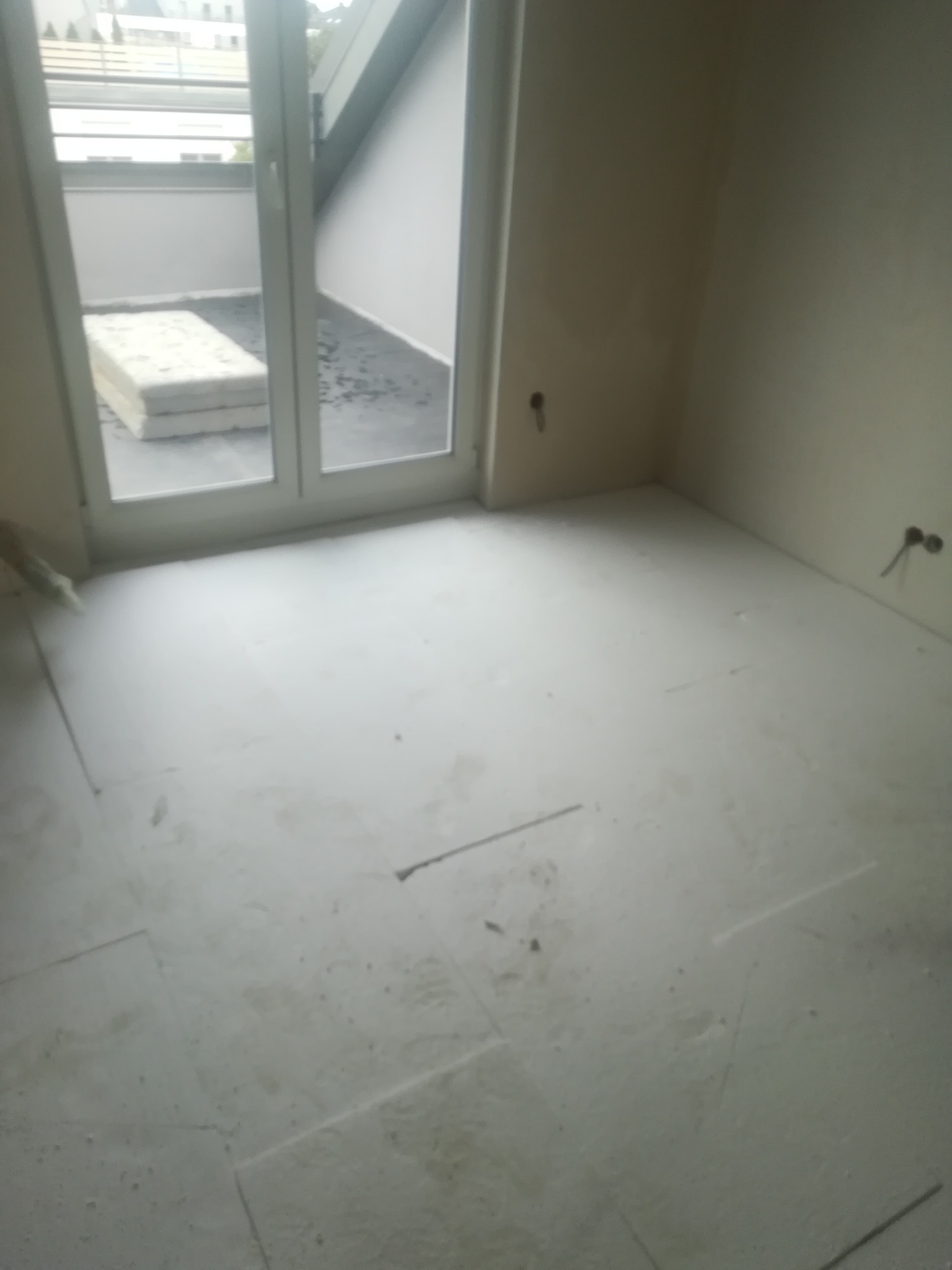 Wnętrze pokoju w trakcie prac wykończeniowych; widoczna wylewka podłogowa z widocznymi szczelinami dylatacyjnymi, okno balkonowe z widokiem na taras z materacem.
