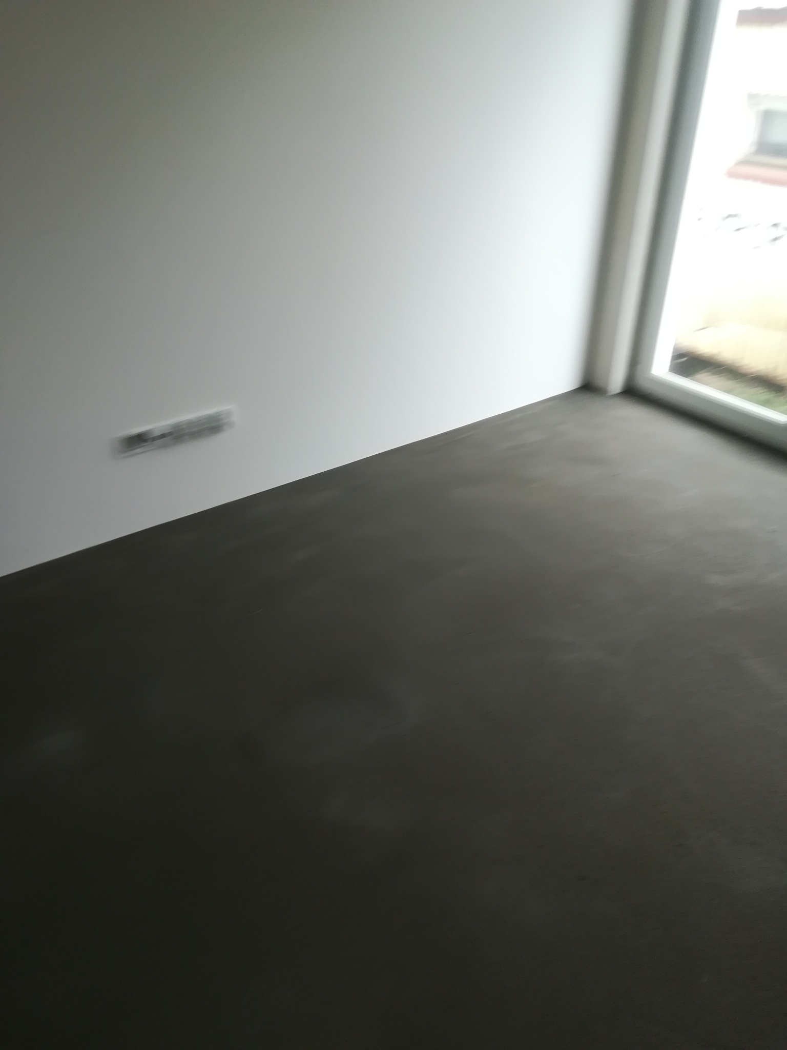 Świeżo wylana, gładka, szara posadzka w rogu pokoju, widoczne gniazdko elektryczne na ścianie i okno z widokiem na zewnątrz.