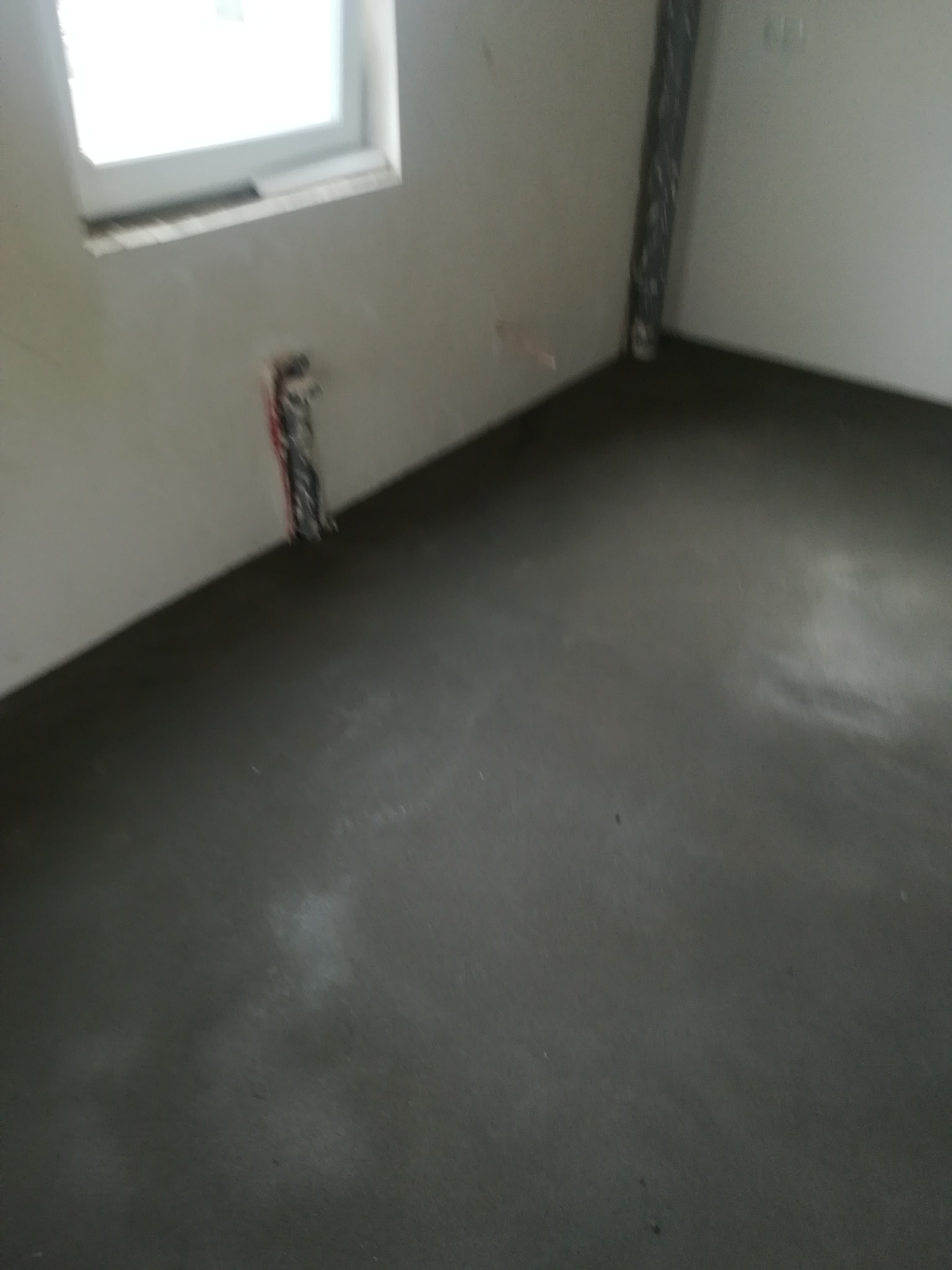 Świeżo wylana, ciemnoszara posadzka w pomieszczeniu z oknem, widoczne kable elektryczne wystające ze ściany.
