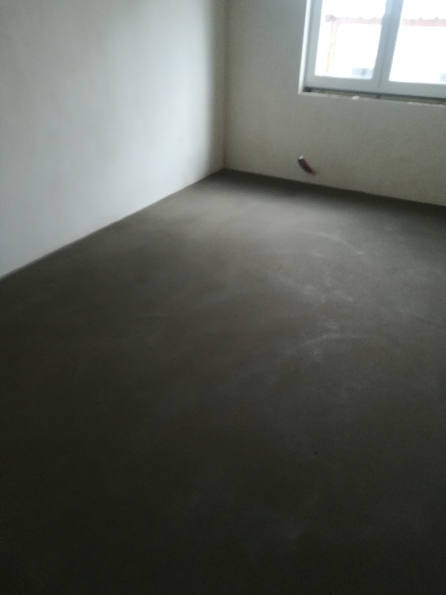 Świeżo wylana, szara posadzka w pustym pokoju z oknem. Widoczne delikatne zacieki i nierówności na powierzchni.