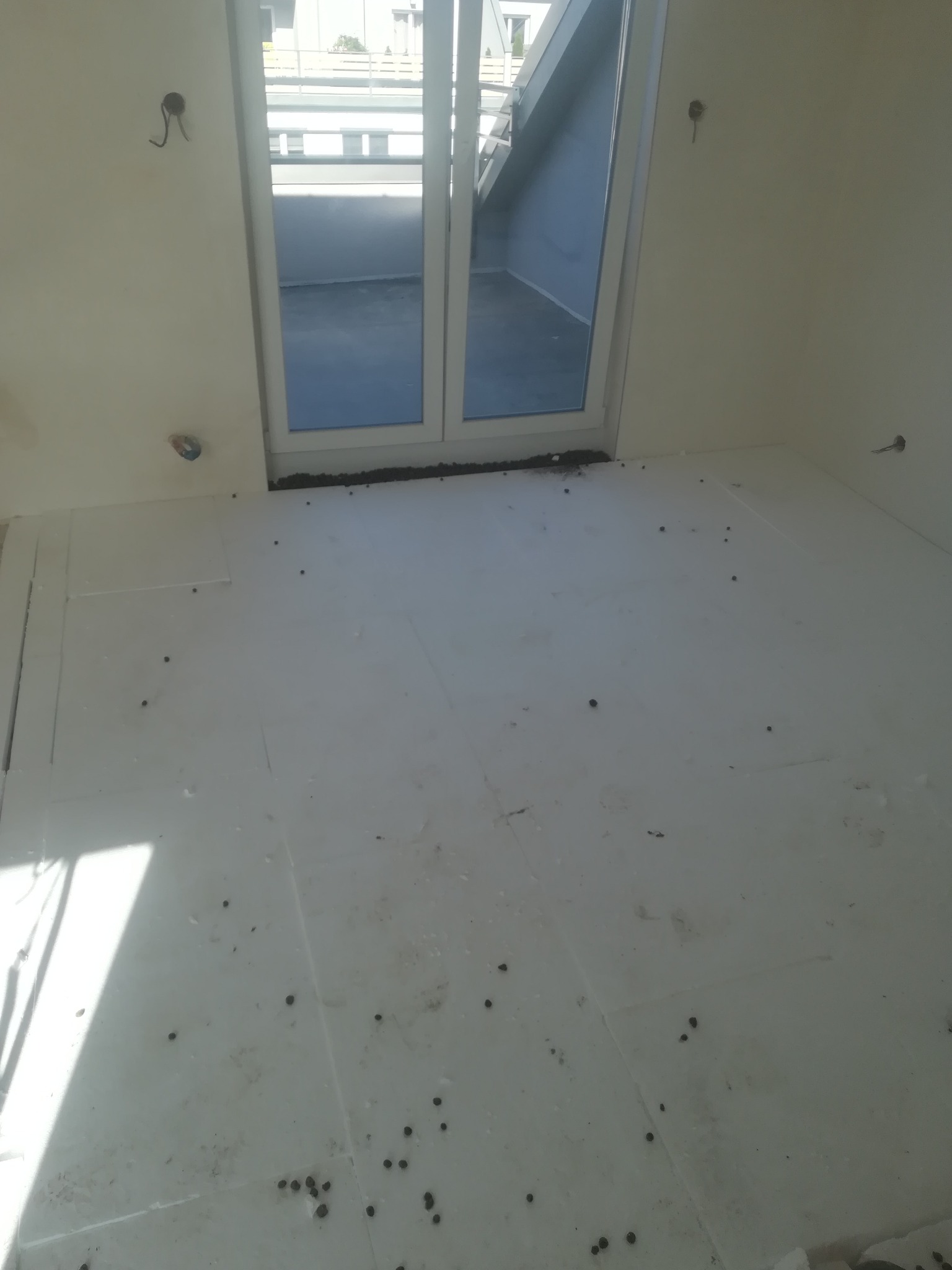 Przygotowanie podłoża pod wylewkę: styropianowe płyty izolacyjne z widocznymi śladami zanieczyszczeń i rozsypanych kulek styropianu, okno balkonowe w tle.