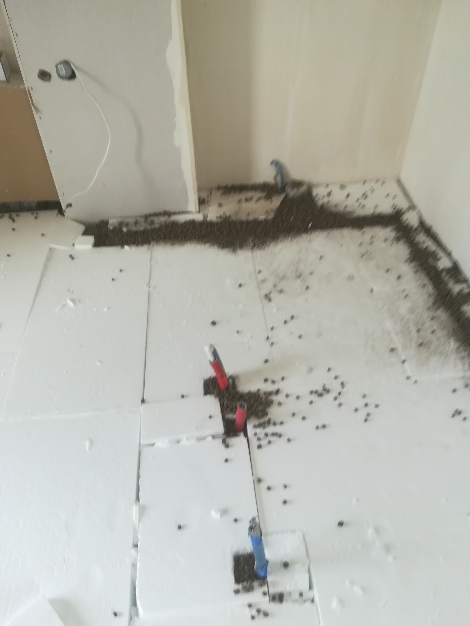 Przygotowanie podłoża: białe płyty izolacyjne rozłożone na podłodze z widocznymi rurami instalacyjnymi i rozsypanym granulatem wyrównującym.