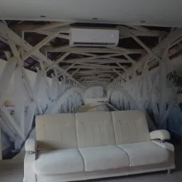 Klimatyzator zamontowany nad fototapetą przedstawiającą drewniany most, widoczny salon z jasną sofą i rośliną doniczkową.