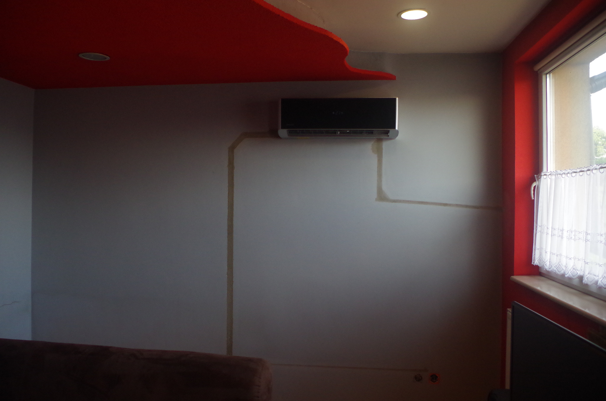 Czarny klimatyzator na szarej ścianie z widocznymi śladami po instalacji przewodów chłodniczych, częściowo ukrytych w ścianie, z czerwonym akcentem na suficie i ramie okna.