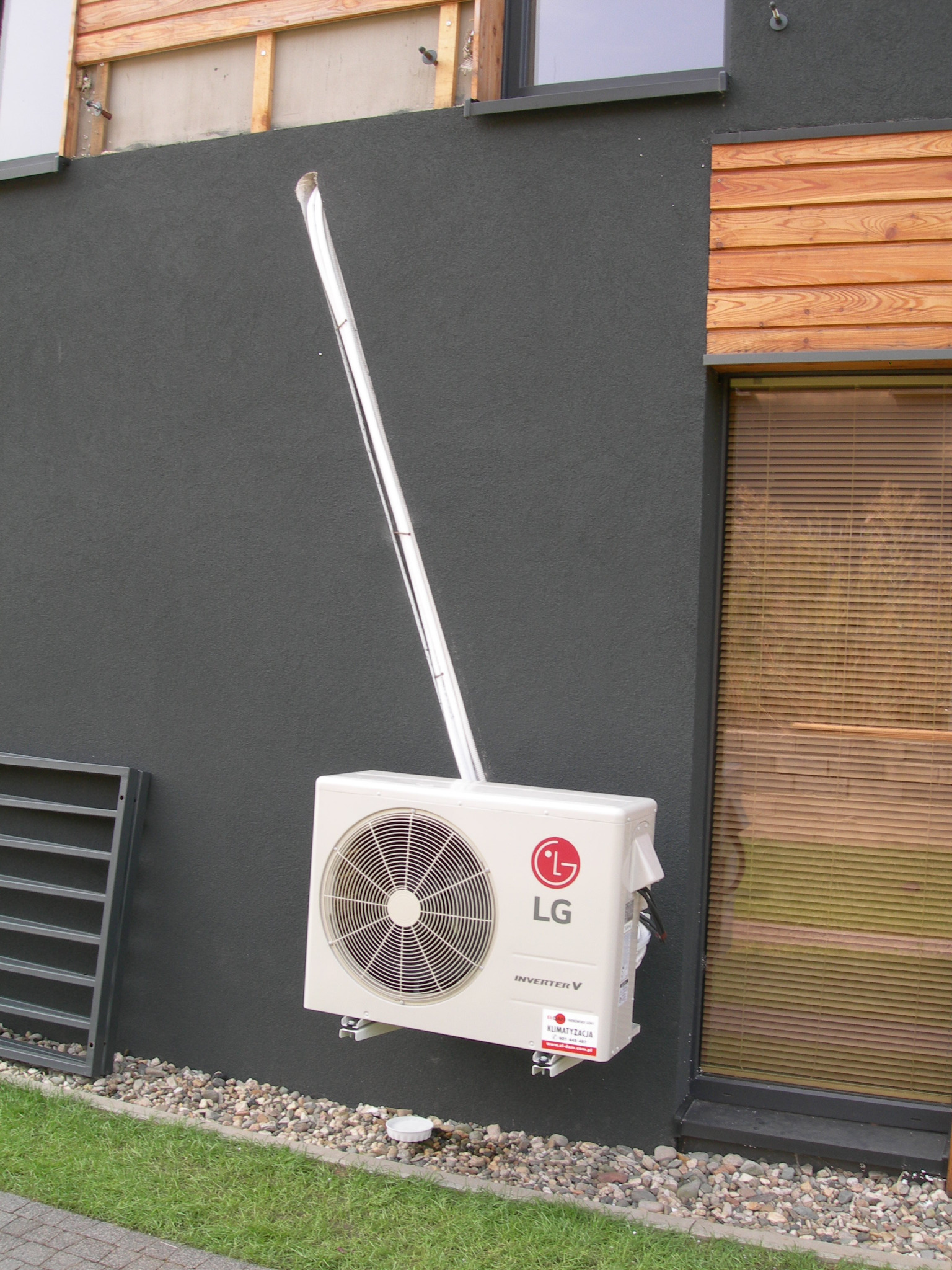 Biała jednostka zewnętrzna klimatyzatora LG Inverter V zamontowana na ciemnoszarej ścianie budynku z drewnianą okładziną i oknem z żaluzjami, widoczna instalacja odprowadzająca skropliny.