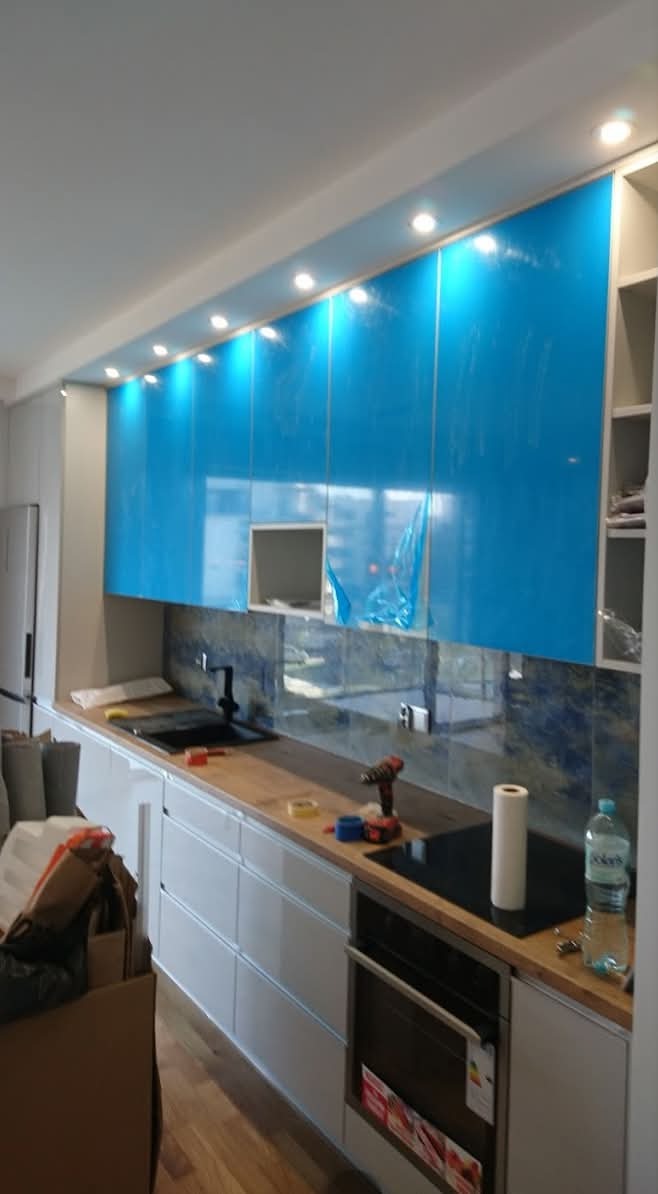 Nowoczesna kuchnia w trakcie montażu: białe szafki dolne, górne z niebieską folią ochronną, drewniany blat, ciemna ściana nad blatem, oświetlenie punktowe.