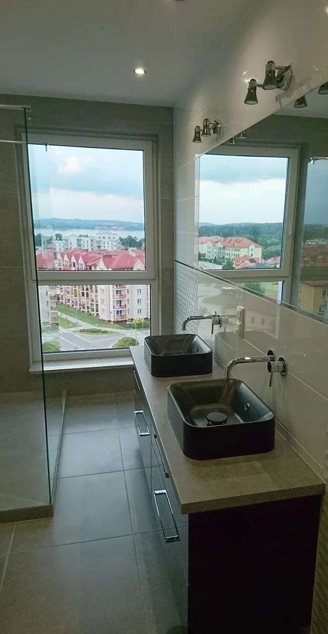 Nowoczesna łazienka z dwoma czarnymi umywalkami na blacie z widokiem na miasto przez duże okno. Minimalistyczny design z szarą kolorystyką.