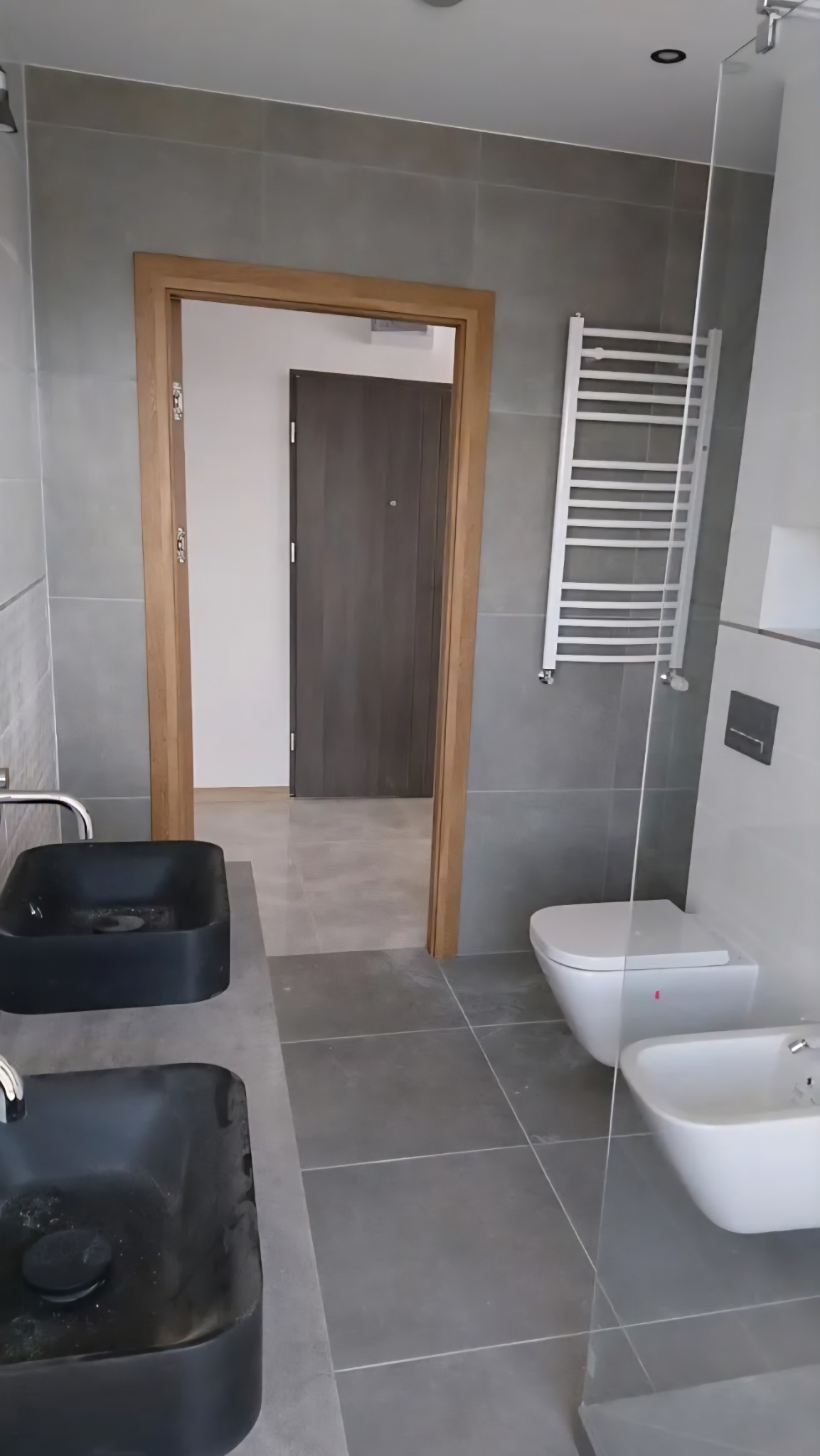 Nowoczesna łazienka z czarnymi umywalkami, szarymi płytkami i grzejnikiem drabinkowym. Widoczny sedes, bidet i drewniane obramowanie drzwi.