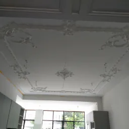 Biały sufit z dekoracyjnymi, gipsowymi ornamentami w stylu retro, widoczne fragmenty ścian i okna w tle.