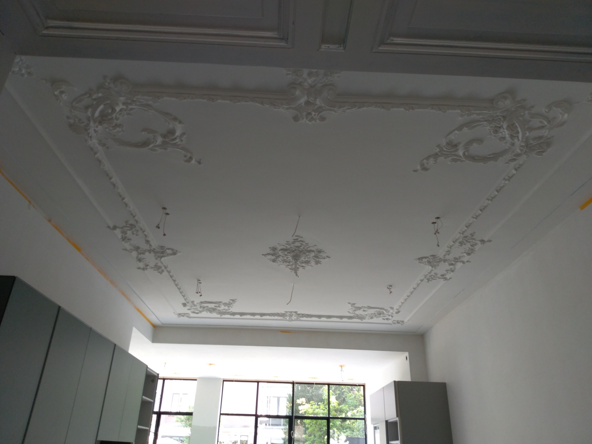 Biały sufit z dekoracyjnymi, gipsowymi ornamentami w stylu retro, widoczne fragmenty ścian i okna w tle.