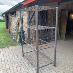 Metalowy regał z czterema półkami z siatki, spawany, surowy metal, na tle ceglanego budynku gospodarczego z otwartymi wrotami.