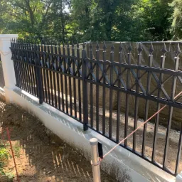 Częściowo zamontowane, czarne, metalowe ogrodzenie z dekoracyjnymi grotami na betonowej podmurówce, widoczne narzędzia i teren budowy w tle.