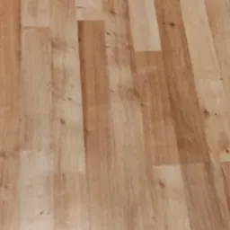 Jasne, drewnopodobne panele podłogowe ułożone wzdłużnie, z widocznymi łączeniami i subtelnym wzorem słojów, lekko rozmyte przez nieostre ujęcie.