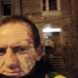 Portret mężczyzny w żółtej kurtce na tle budynku z oknami i schodami, prawdopodobnie wykonany nocą.