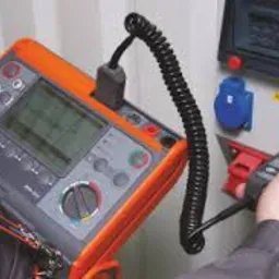 Miernik elektryczny w pomarańczowej obudowie podłączony do gniazda w rozdzielnicy, widoczna dłoń osoby wykonującej pomiar, czarny przewód spiralny.