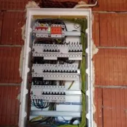 Rozdzielnica elektryczna z zamontowanymi bezpiecznikami i przewodami, widoczna na tle ściany z czerwonej cegły.