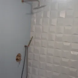 Bateria prysznicowa w kolorze złotym zamontowana na ścianie z trójwymiarowych, białych płytek w wyremontowanej łazience nad białą wanną.