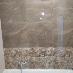 Wykończona ściana nad wanną z brązowych płytek imitujących marmur u góry i płytek z motywem kwiatowym poniżej, widoczna bateria wannowa.