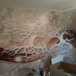 Ściana z fototapetą przedstawiającą stylizowane drzewa w kształcie profilów twarzy, w ciepłej tonacji brązowo-beżowej z efektem bokeh, widoczne kartony i akcesoria montażowe.