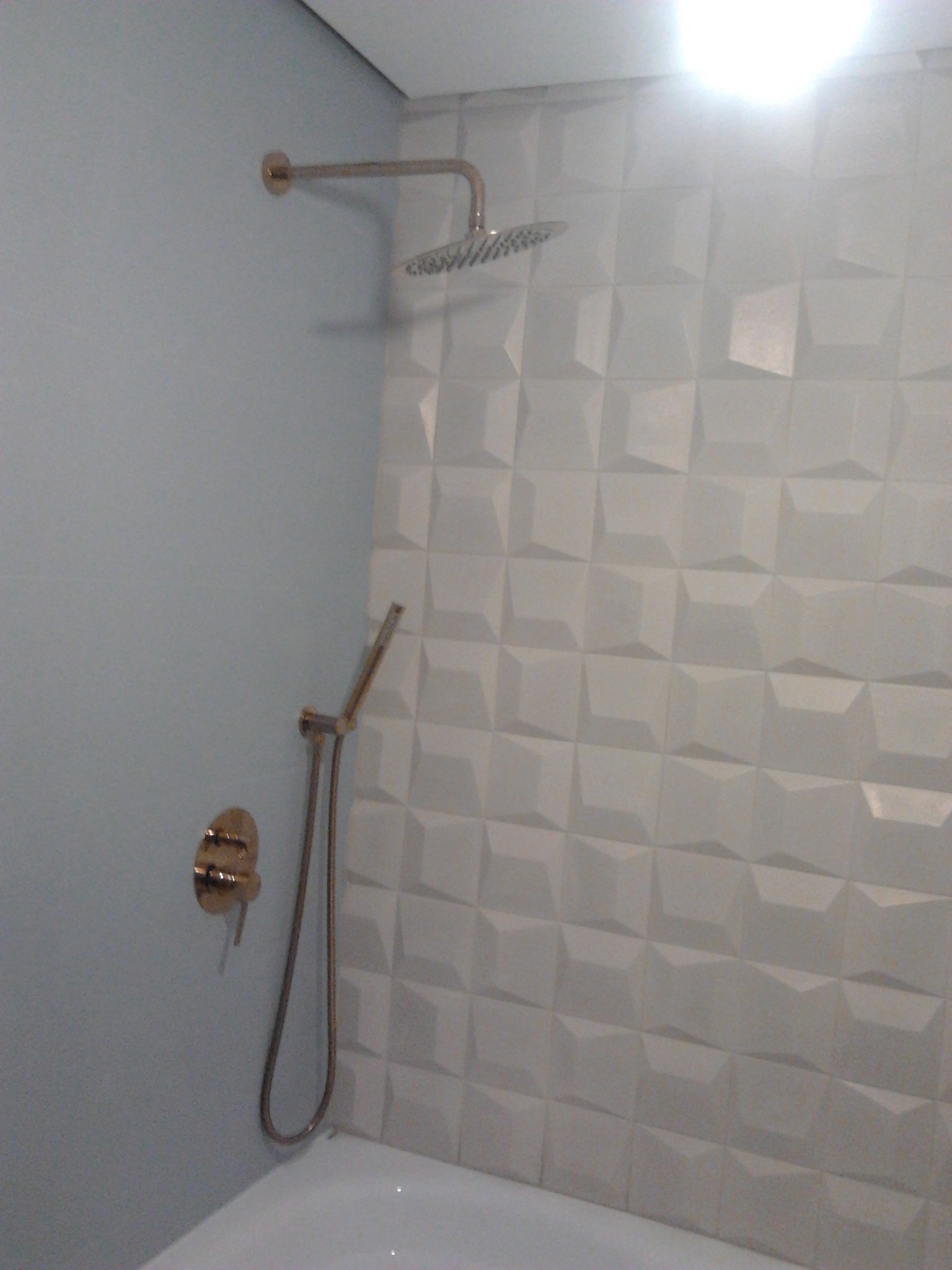 Bateria prysznicowa w kolorze złotym zamontowana na ścianie z trójwymiarowych, białych płytek w wyremontowanej łazience nad białą wanną.