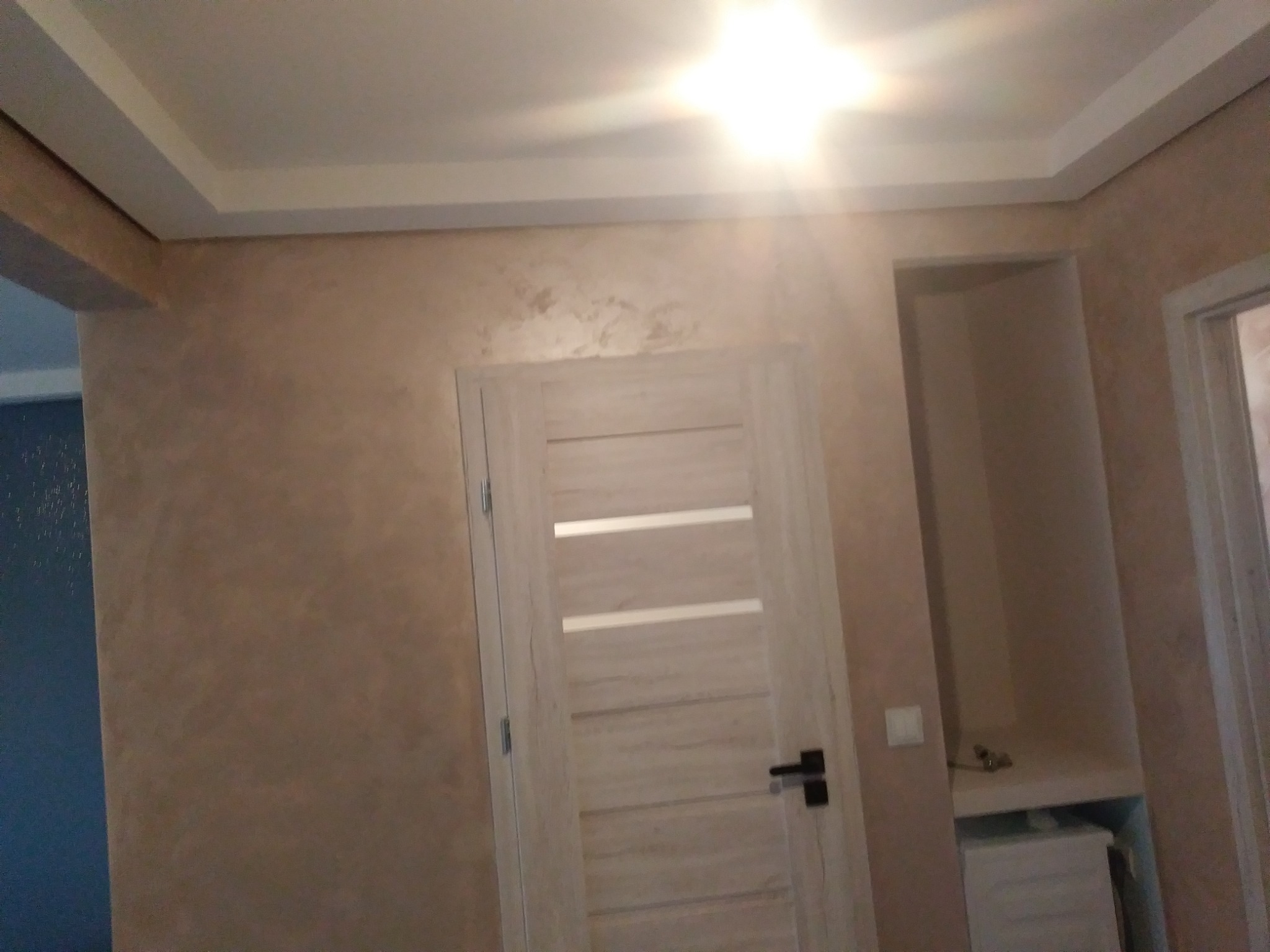 Ściana wykończona dekoracyjnym tynkiem w odcieniach beżu, z widocznymi nieregularnymi wzorami, obok wewnętrzna drewniana drzwi z poziomymi przeszkleniami i czarną klamką, w tle wnęka z szafką.