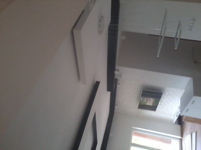 Fragment wnętrza: biała ściana z ramkami i czarnym elementem dekoracyjnym, widoczny fragment okna i telewizora na ścianie z fakturą. Ujęcie z dołu.